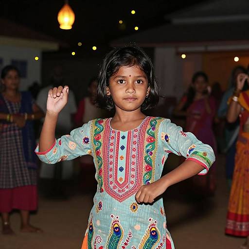 Photograph of a young Indian girl with dark hair, wearing a light blue, embroidered dress, raising her right hand, standing in a dimly lit