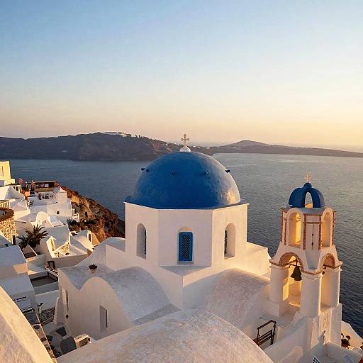 Photograph of a Greek island sunset with white-washed buildings, blue domes, and a church bell tower overlooking a calm sea.