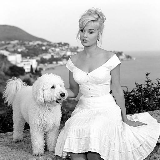 Black-and-white photograph of a blonde woman in an off-shoulder white dress, standing beside a fluffy white dog, overlooking a coastal town.