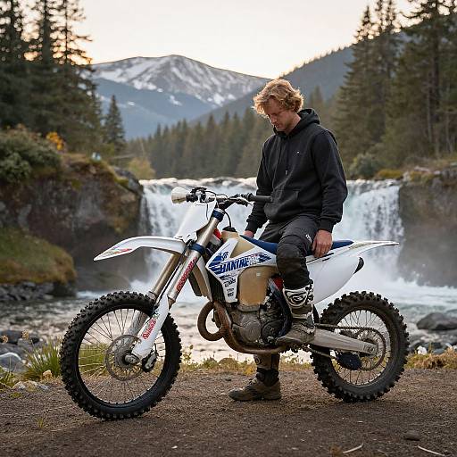 Photograph of a blonde man in a black hoodie and pants, sitting on a white motocross bike beside a waterfall in a forested mountain area