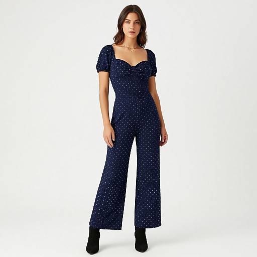 Photograph of a woman with medium-length brown hair, wearing a dark blue, sparkling, short-sleeve jumpsuit and black heels, standing against