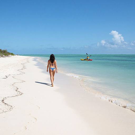 Photograph of a woman in a blue bikini walking on a white sandy beach, with a kayaker in the turquoise ocean under a clear blue sky.