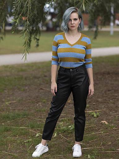 Photograph of a white woman with short, blue-gray hair, wearing a blue and yellow striped sweater, black pants, and white sneakers, standing in
