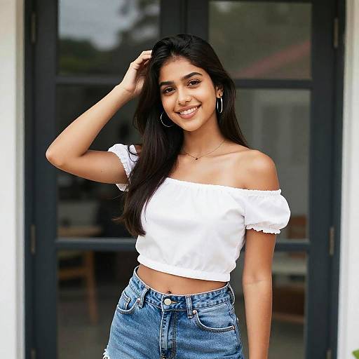 Photograph of a smiling young woman with long dark hair, wearing a white off-shoulder top and blue jeans, standing in front of a glass