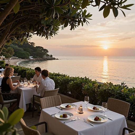 Photograph of a romantic seaside restaurant at sunset, with diners enjoying meals on white-clothed tables under leafy overhangs, overlooking a