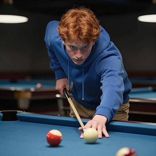 Photograph of a red-haired man in a blue hoodie, intensely aiming a pool cue at a white ball on a blue pool table.