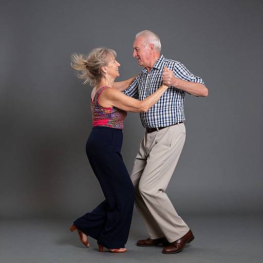 Photograph of an elderly white couple dancing, wearing checkered shirt and floral tank top, black pants, and red shoes, against a gray background.