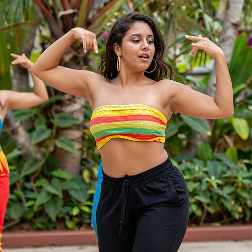 Photograph of a curvy Latina woman with medium skin tone, wearing a yellow-green-red striped tube top and black pants, dancing outdoors with lush green