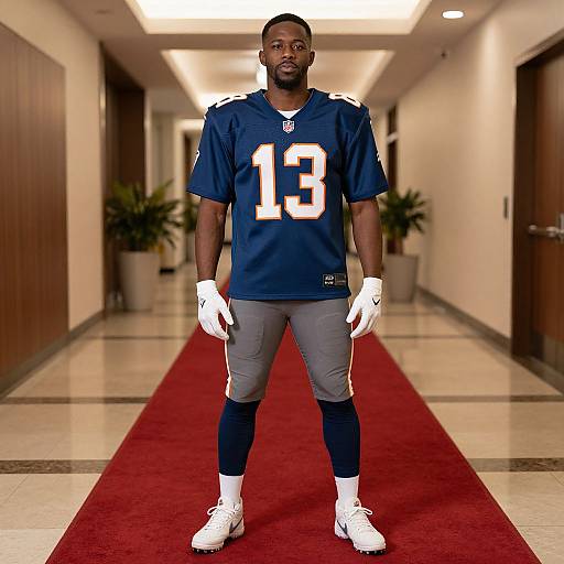 Photograph of an African-American male football player in a blue #13 jersey, gray pants, white gloves, and white cleats, standing on a