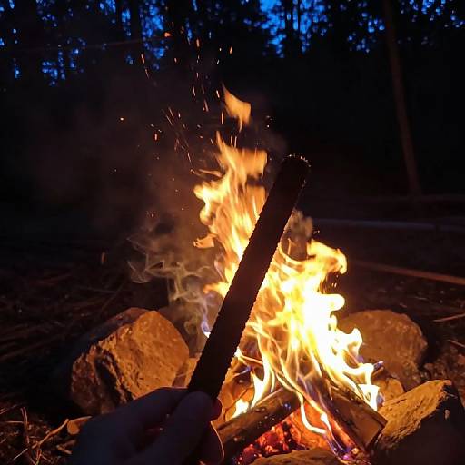 Photograph of a vibrant campfire at night, with bright orange flames, glowing embers, and smoke, surrounded by large rocks and wooden logs.
