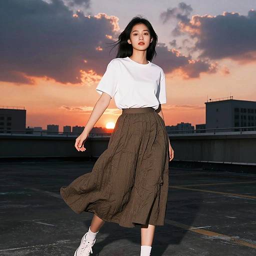 Photograph of an Asian woman with long black hair, wearing a white t-shirt and brown pleated skirt, walking on a rooftop at sunset, orange