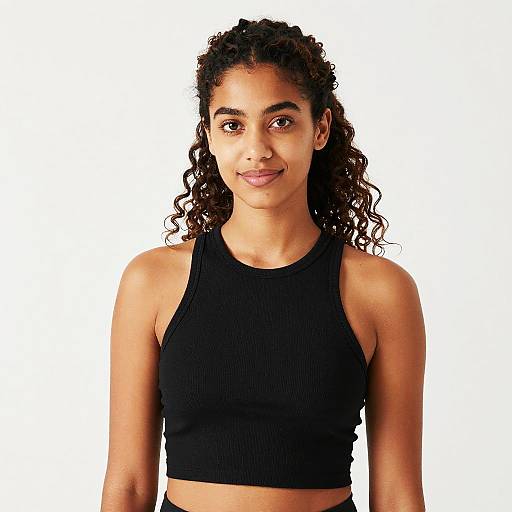 Photograph of a young woman with curly black hair, brown skin, and brown eyes, wearing a black ribbed crop top, smiling against a plain