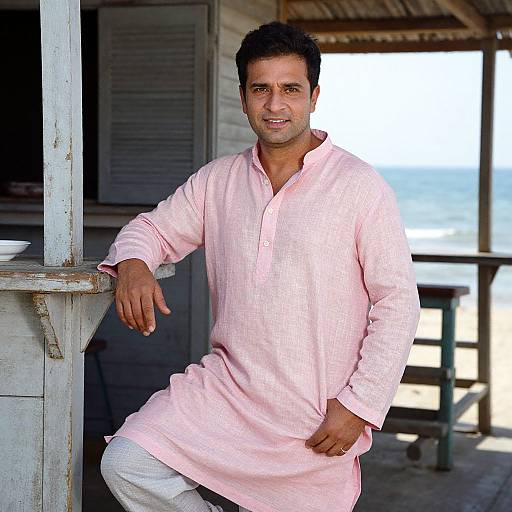 Photograph of a smiling, dark-haired man in a pink, long-sleeve, checkered kurta, leaning against a wooden structure with a