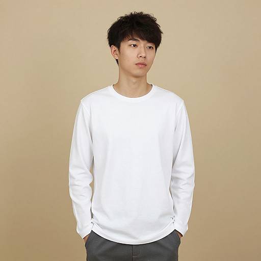 Photograph of an Asian man with short black hair, wearing a plain white long-sleeve shirt and dark pants, standing against a beige background,