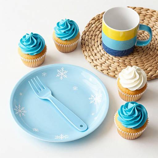 Photograph of blue and white frosted cupcakes, yellow-blue striped cup on woven mat, light blue snowflake plate with fork.