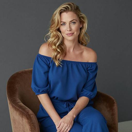 Photograph of a blonde woman with wavy hair, wearing an off-shoulder royal blue top and matching pants, seated in a brown velvet chair