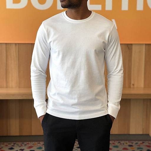 Photograph of a black man with short hair, wearing a plain white long-sleeve shirt and black pants, standing against a wooden wall with an
