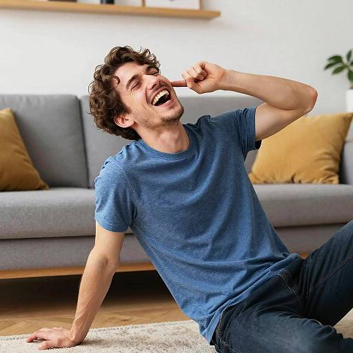 Photograph of a smiling, curly-haired man in a blue t-shirt and jeans, sitting on a wooden floor, pointing at his ear with a playful