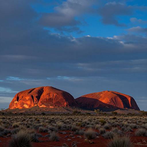 Photograph of two large, red-orange rock formations bathed in sunlight, set against a blue and gray clouded sky in a desert landscape with sparse