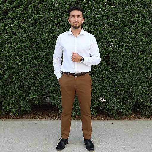 Photograph of a handsome man with short black hair, beard, wearing a white shirt, brown pants, and black shoes, standing in front of a
