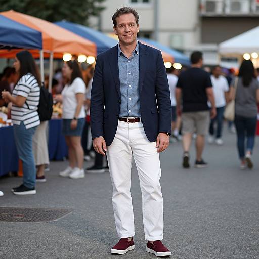 Photograph of a smiling middle-aged man in a navy blazer, blue shirt, white pants, and maroon sneakers, standing at an outdoor market