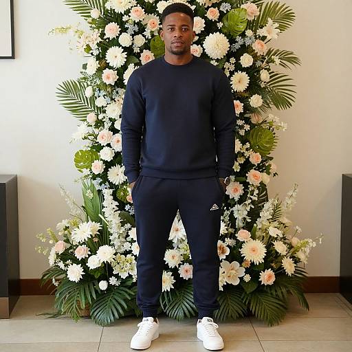 Photograph of a tall, muscular Black man in a black sweatshirt and pants, standing in front of a vibrant floral arrangement with white and pink flowers