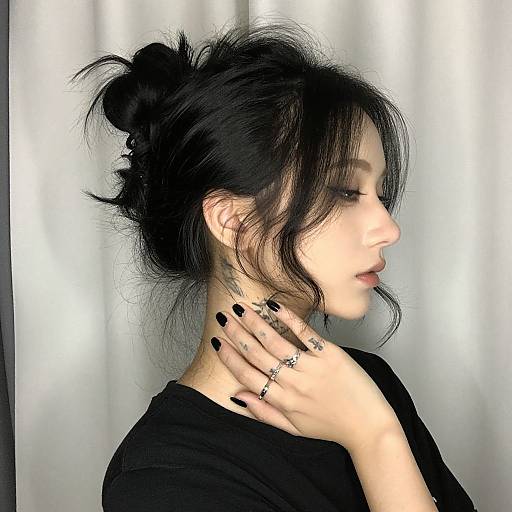 Photograph of an Asian woman with black hair in a messy bun, black nails, black top, and multiple rings, looking to the side against a