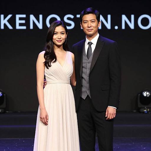 Photograph of an Asian couple on a fashion runway; she wears a white, sleeveless, pleated dress, he in a black suit with gray
