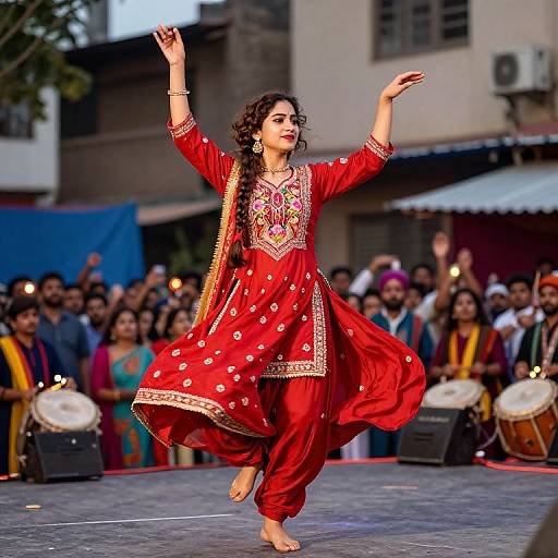 Photograph of a young South Asian woman in a vibrant red traditional outfit, dancing barefoot with arms raised, in front of a cheering crowd and musicians