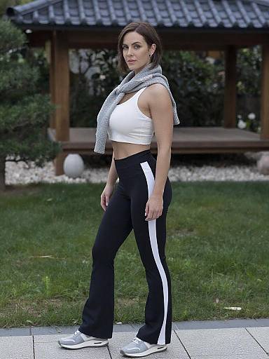 Photograph of a fit woman with medium brown hair, wearing a white sleeveless top, black track pants with white stripes, gray scarf, and white