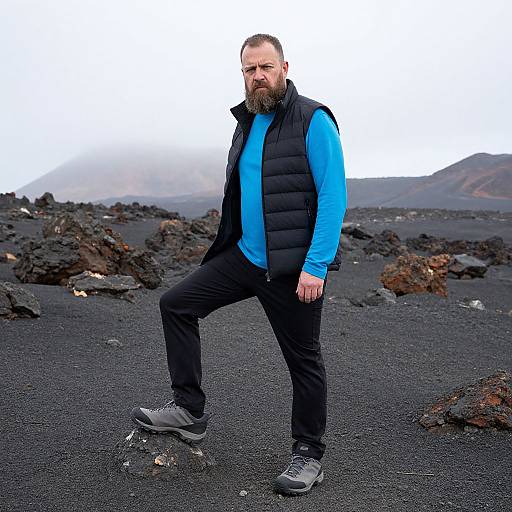 Photograph of a bearded man in a blue shirt, black puffer vest, and gray shoes, standing confidently on a black volcanic landscape with rocky