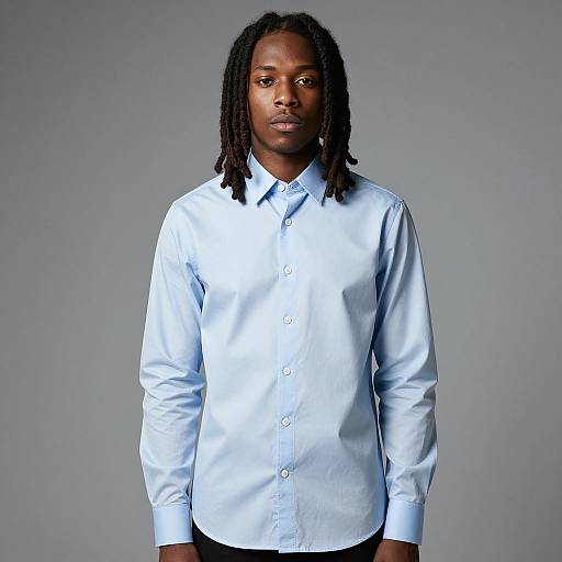 Photograph of a Black man with medium brown skin, shoulder-length dreadlocks, wearing a light blue button-up shirt, standing against a plain gray background