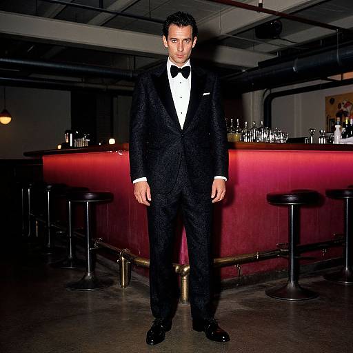 Photograph of a handsome man in a black tuxedo with a bow tie, standing in a dimly lit bar with red-lit counter and