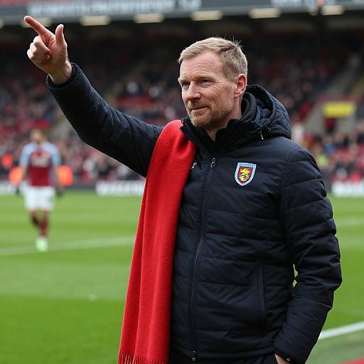 Photograph of a bearded male football coach in a black puffer jacket with a red scarf, pointing, in a stadium with blurred players in the
