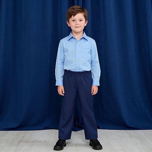 Photograph of a young boy with short brown hair, wearing a light blue button-up shirt, dark navy pants, and black shoes, standing against a