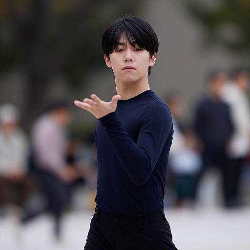 Photograph of a young Asian man with short black hair, wearing a black long-sleeve shirt, gesturing with his right hand against a blurred