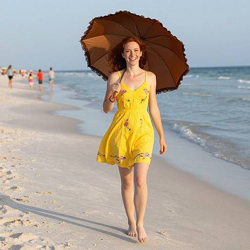 Photograph of a smiling young woman with curly red hair, wearing a yellow floral dress and holding a brown umbrella, walking barefoot on a sunny beach