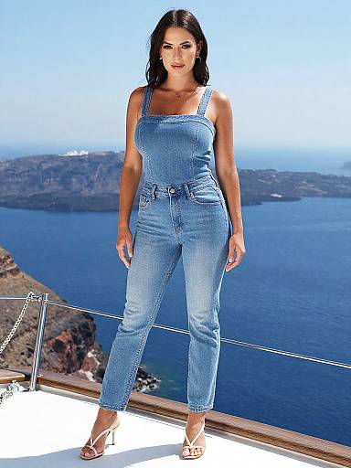 Photograph of a brunette woman in blue denim overalls and white sandals, standing on a balcony overlooking a blue sea and mountainous landscape.