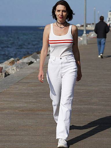 Photograph of a woman with short black hair, wearing a white tank top with red stripes, white pants, and white shoes, walking on a wooden