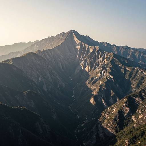 Photograph of a rugged mountain peak illuminated by sunlight, casting shadows over dark, forested valleys and winding trails, under a clear blue sky.
