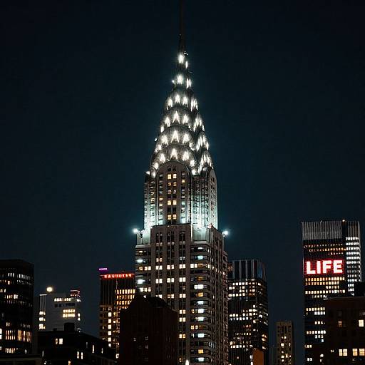 Photograph of a cityscape at night, featuring a brightly lit, ornate skyscraper with illuminated spires, surrounded by other lit buildings, including