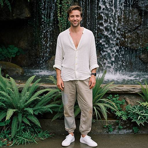 Photograph of a smiling bearded man with light brown hair, wearing a white button-up shirt, beige pants, and white sneakers, standing in front