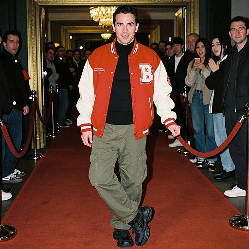 Photograph of a man in a red and white letterman jacket, black shirt, and green cargo pants, walking down a red carpet with a crowd