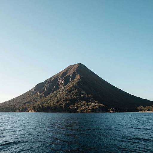 Photograph of a solitary, triangular, forested mountain rising from a calm, blue ocean under a clear, bright sky.