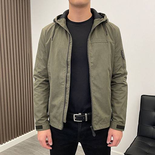 Photograph of a man from shoulders to waist, wearing an olive green jacket, black shirt, and black pants with a belt, standing in front of