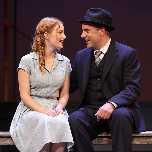Photograph of a smiling couple on stage; woman with red hair in white dress, man in black suit, tie, and hat.
