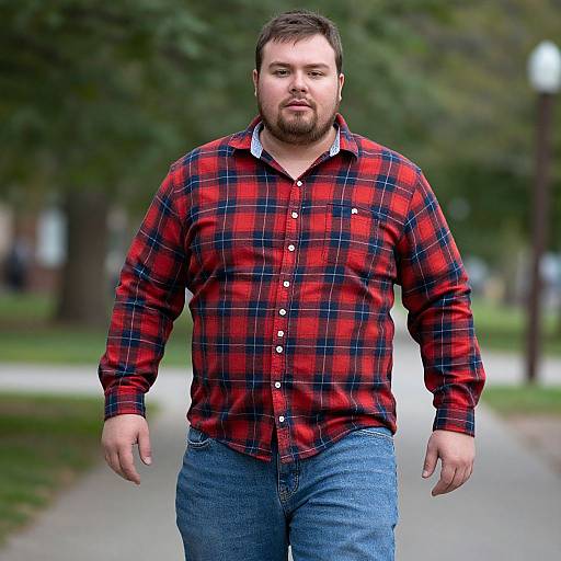 Photograph of a heavyset man with a beard, wearing a red and black plaid shirt and blue jeans, walking on a park path. Bl