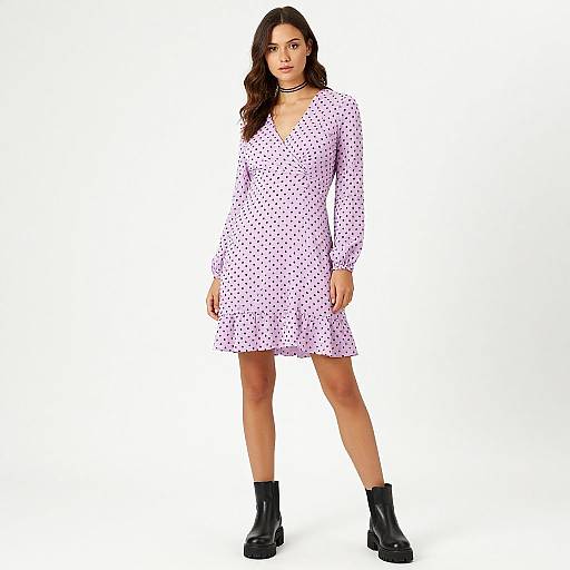 Photograph of a young woman with wavy dark hair, wearing a white polka dot dress, black ankle boots, and a black choker,