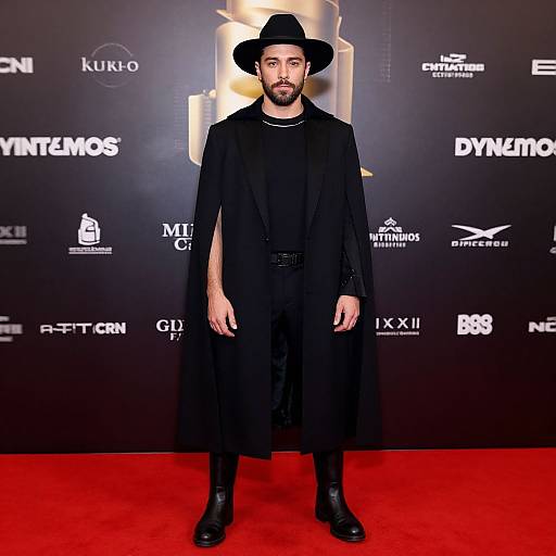 Photograph of a bearded man with olive skin wearing a black hat, long black coat, and boots, standing on a red carpet against a black
