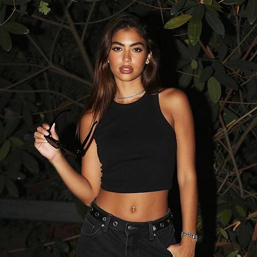Photograph of a young woman with dark brown hair, wearing a black crop top, black pants, and jewelry, standing in front of dark foliage at